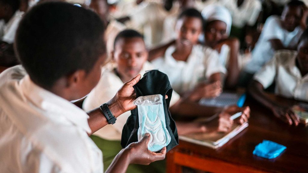 Olan takaa kuvattu tyttö esittelee kuukautissuojaa luokassa istuville muille oppilaille Ugandassa.