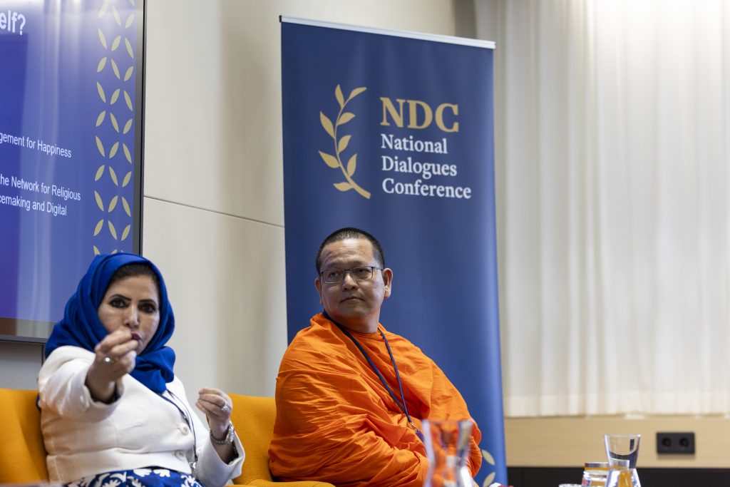 A woman and a man sitting in a room. The woman is talking and pointing to the camera. The man, who is wearing the orange robes of a monk sits beside her and listens. Behind them both is the logo of the National Dialogues Conference