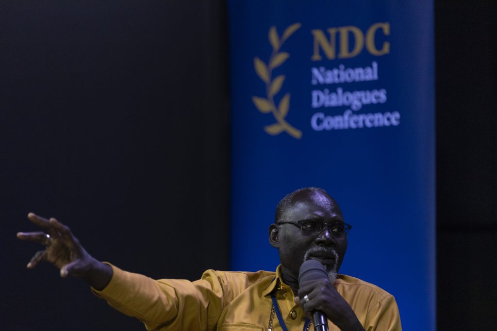 A man with a microphone is talking. Behind him is the logo of the National Dialogues Conference