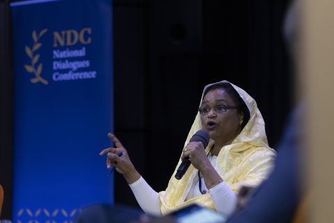 A woman with a microphone is talking. Behind her is the logo of the National Dialogues Conference
