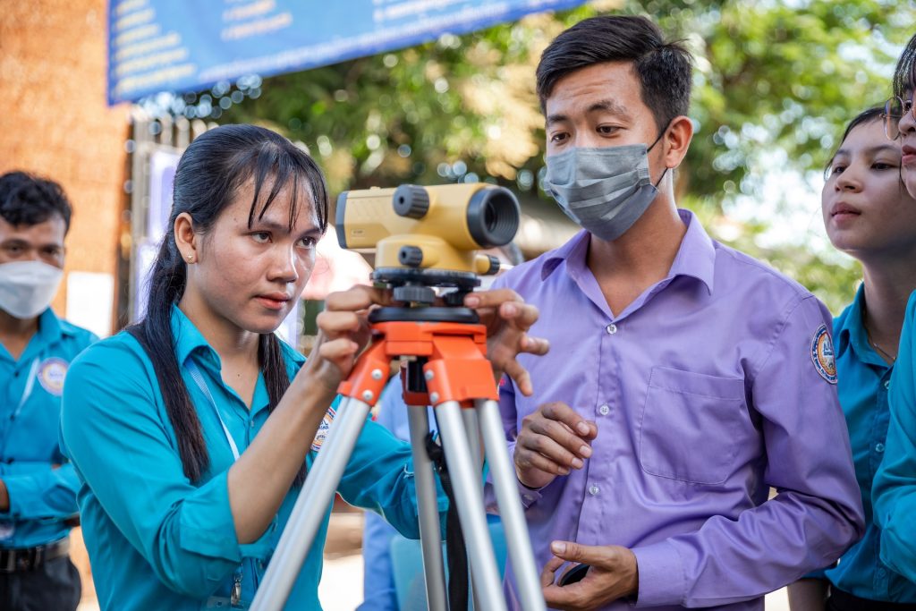 A woman operates a land surveyor's instrument, while a man in an mask looks on
