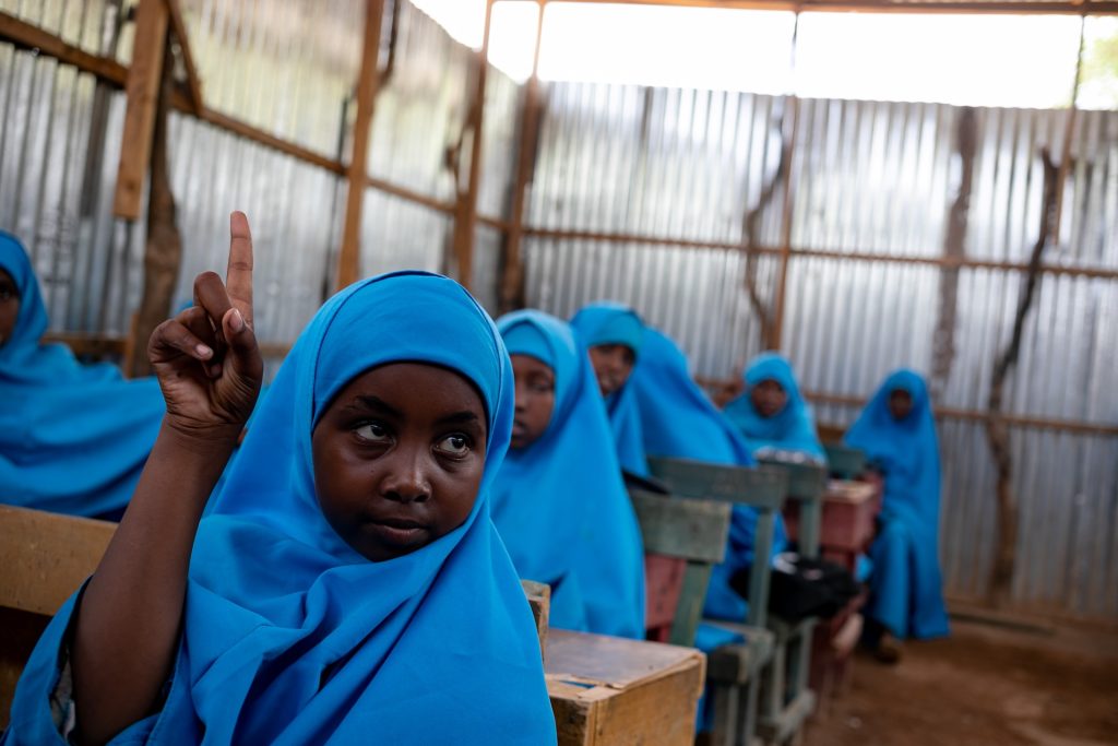 A schoolgirl in a blue hijab puts up her hand in a classroom