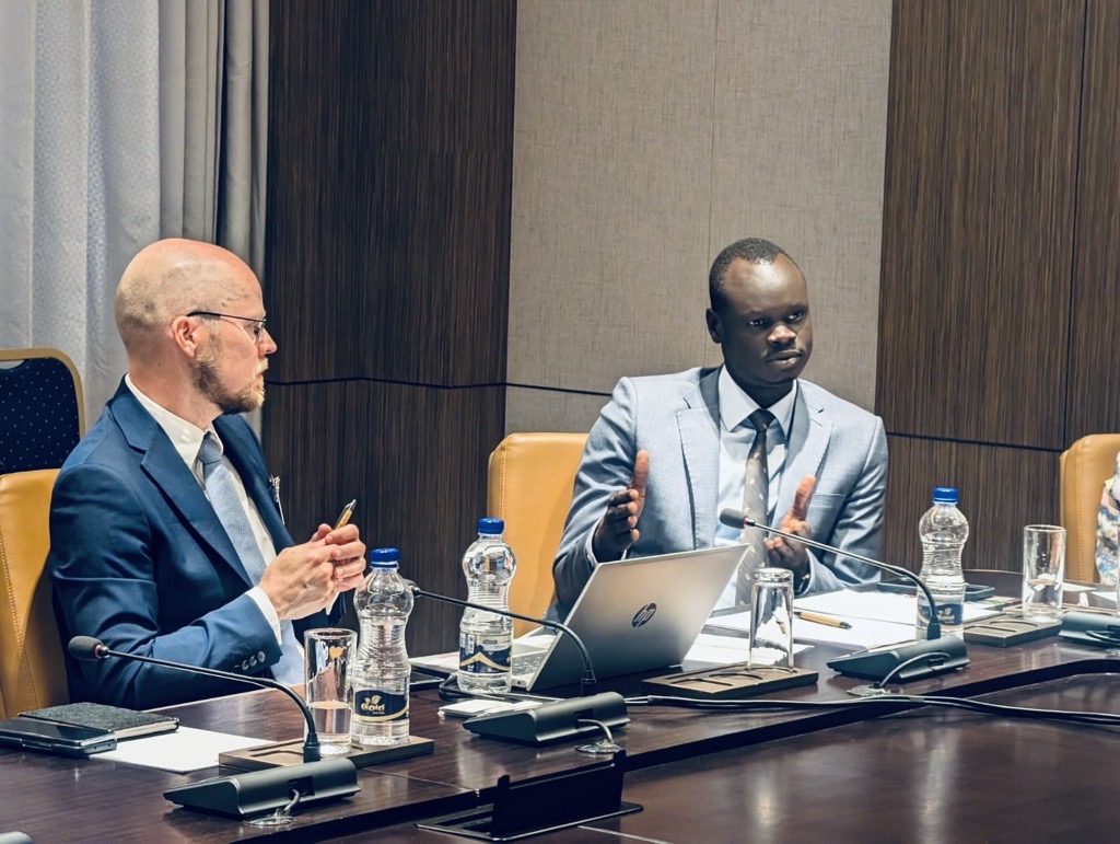 Two men in business suits sit at a conference table indoors. The man on the left is listen as the man on the right gesticulates.