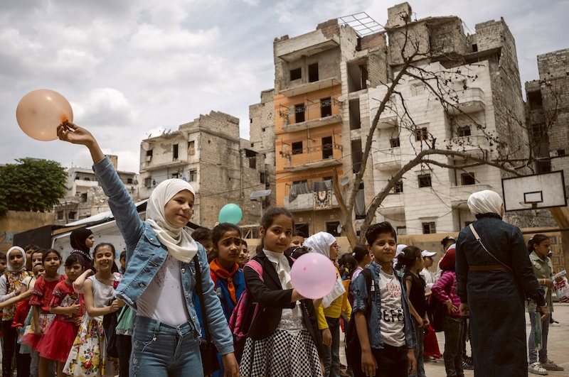 A girl in a hijab holds up a balloon in a playground full of children. In the background are earthquake-damaged buildings.