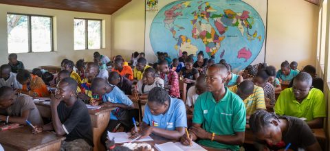 A crowded classroom where many students are listening or writing. In the background there is a large map of the world on the wall.