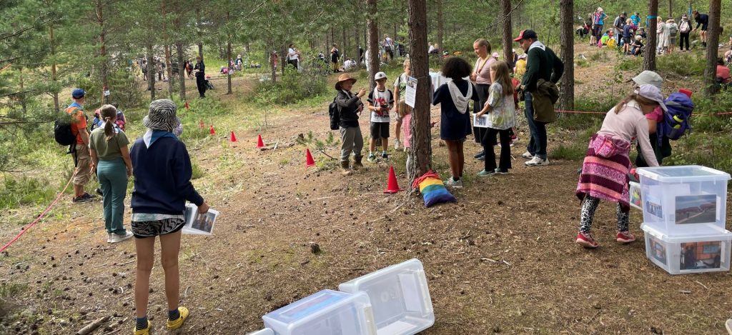 Lapsia metsässä jonossa ja piirimuodostelmassa. Kahdella lapsista on käsissään laatikot. Heidän vieressään on lisää laatikoita sekä punaisia kartioita.