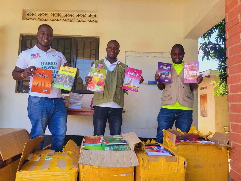 Three men standing in an outside corridor stand in front of several boxes of books. They are smiling at the camera and holding up textbooks in different subjects.