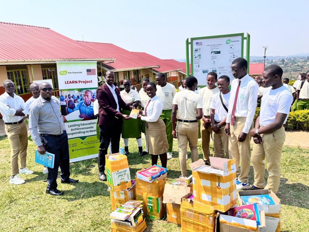 Several adults and young people are standing outside a block of classrooms. Boxes of textbooks are on the ground. In the centre, a man hands a textbook to a schoolgirl. They smile at the camera. FCA and US Government logos are in the background.