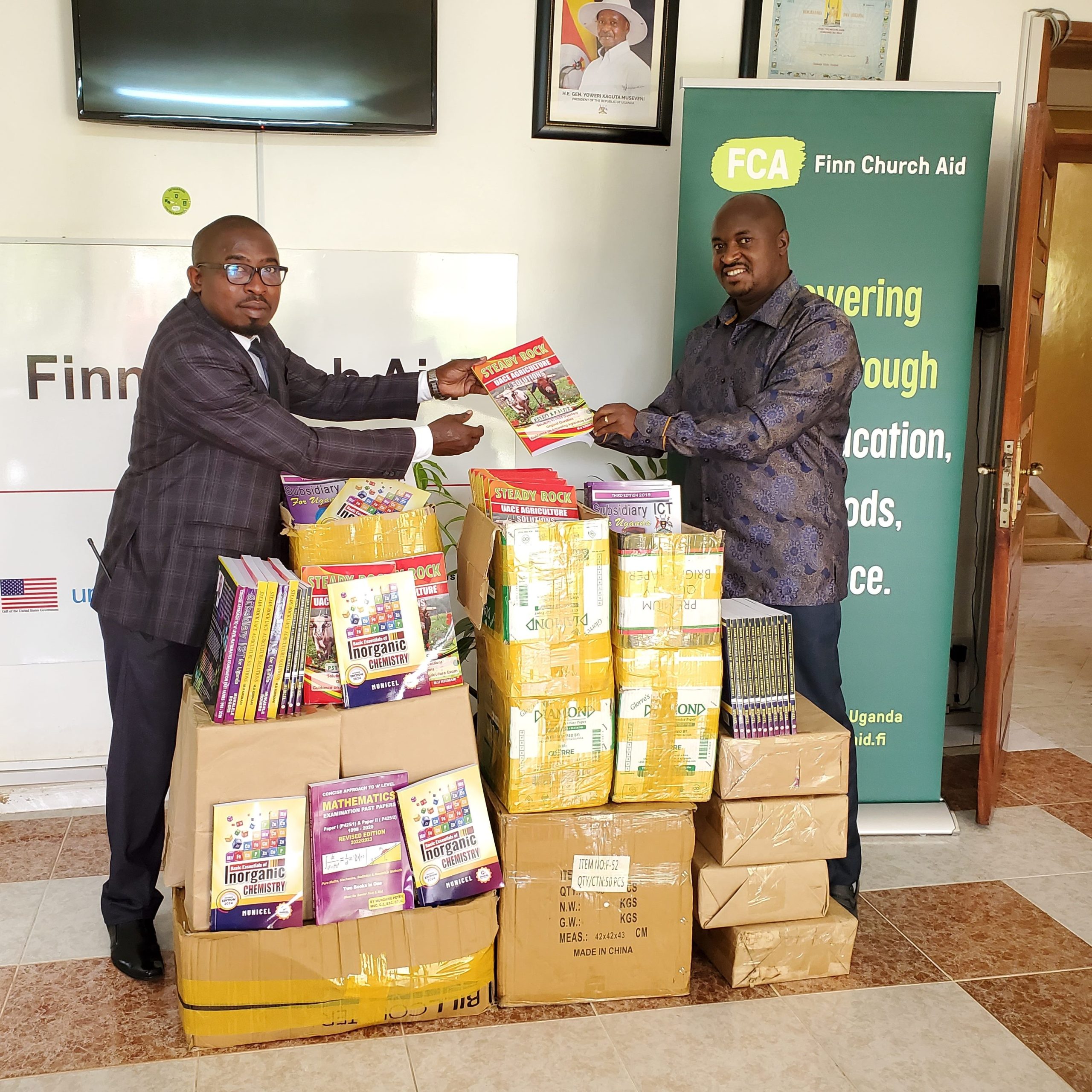 Two men pass books to each other over a pile of boxes. They are both looking at the camera. FCA's logos are in the background.