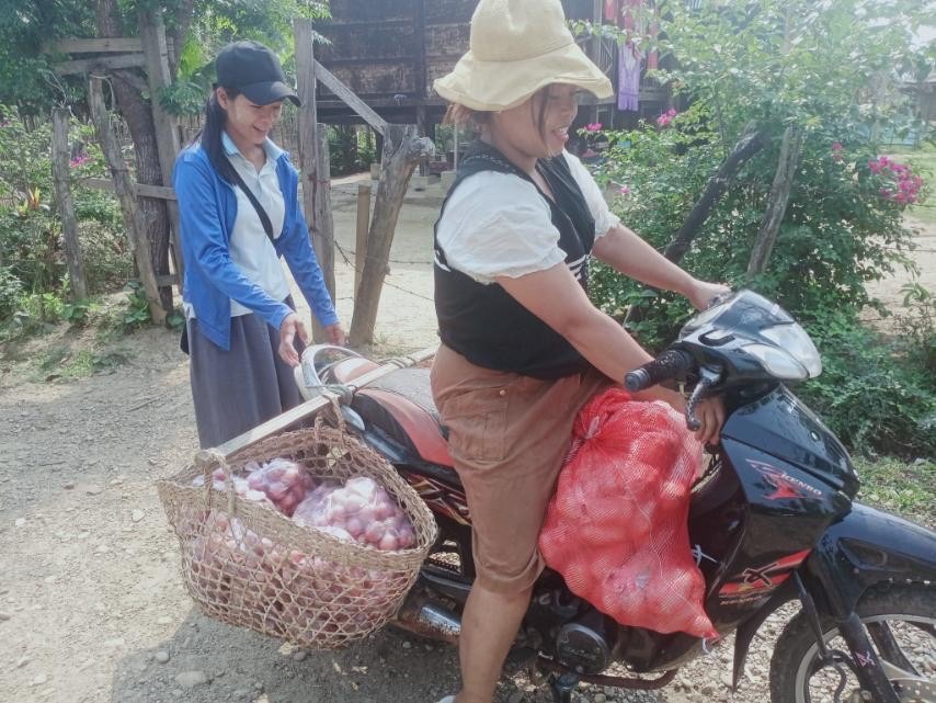 A woman is on a moped outside, which is laden with baskets and bags of onions. Another woman is behind her.