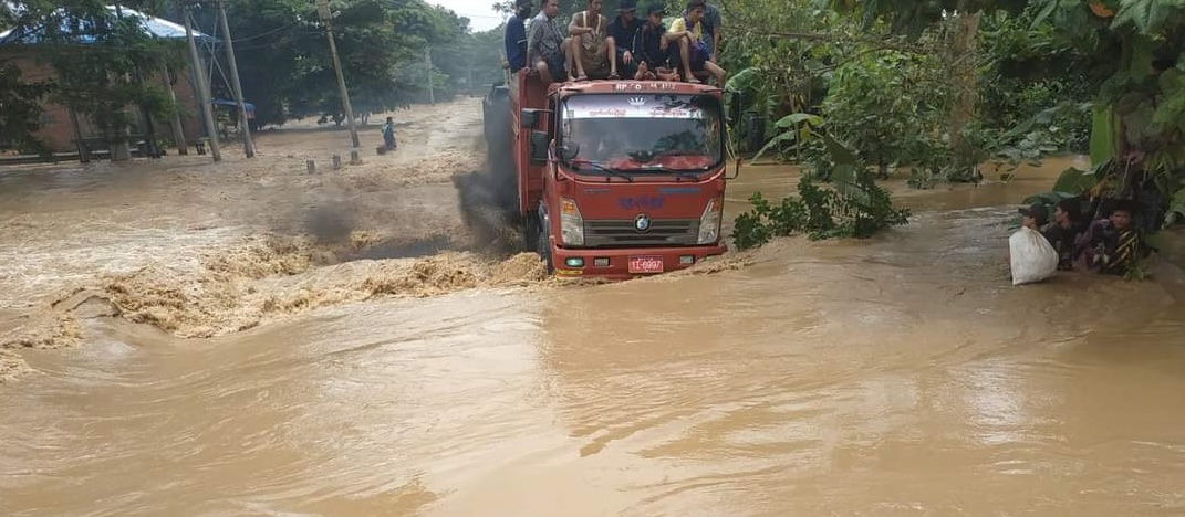 A truck with people sitting on the driver's cab drives through a severely flooded street