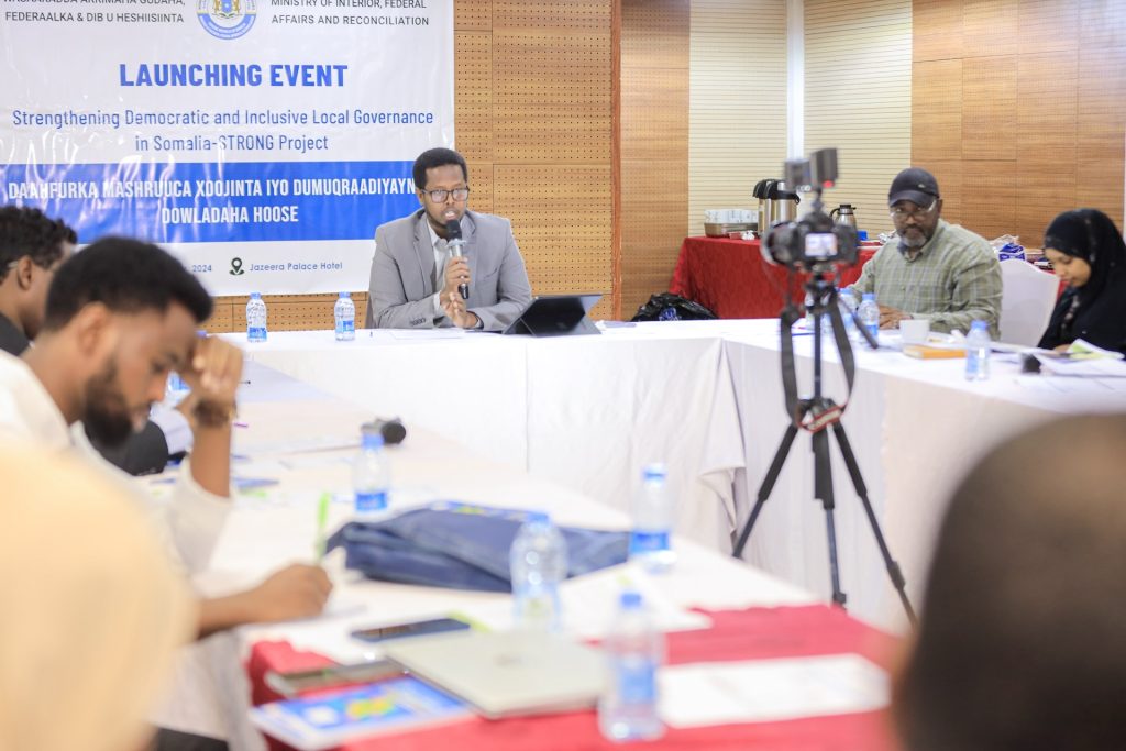 A man speaks into a microphone at a table, while others sitting around listen. A video camera is set up on a tripod recording the speaking man. Behind him there is a banner reading "Launching Event. Strengthening Democratic and INclusive Local Governance in Somalia - STRONG Project"