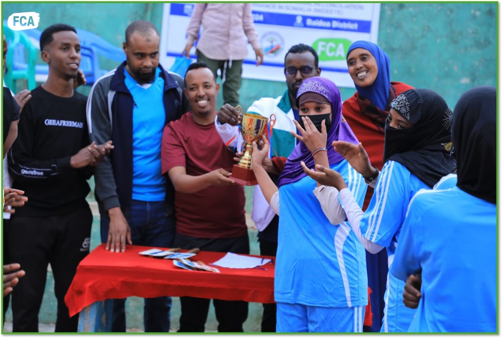 A woman in a hijab wearing a facemask and in sports clothing receives a sports trophy. Men and women standing around her celebrate.