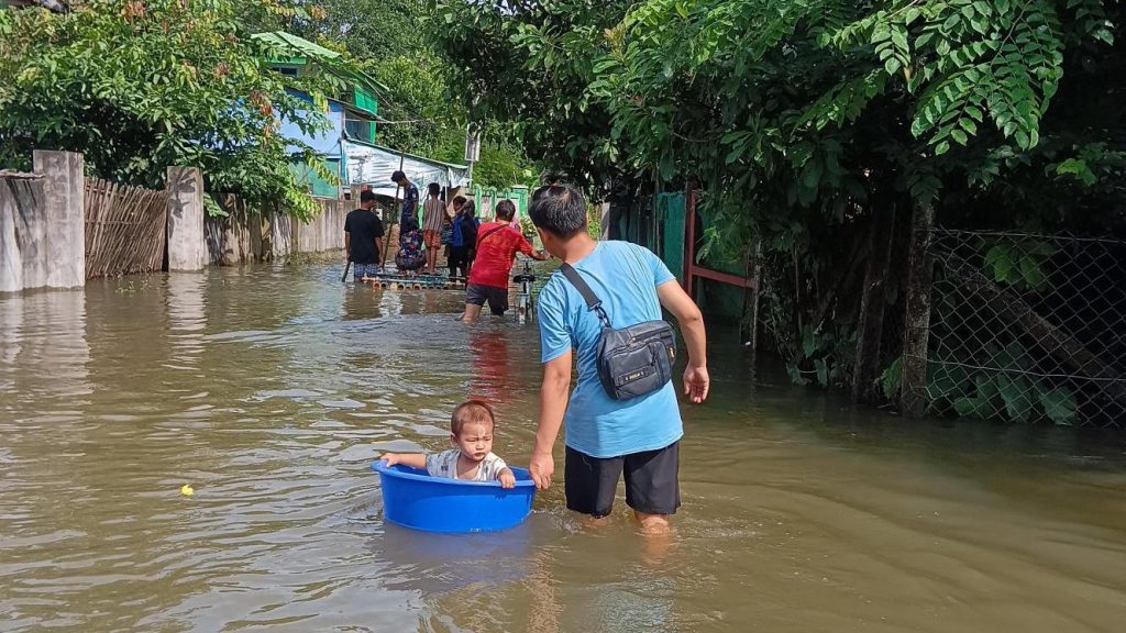 A man pushes a baby in a washing up bowl through a flooded street. 