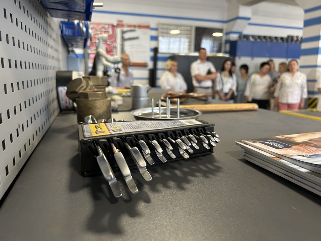 A close up of a number of wrenches on a table. In the background people stand listening in a classroom setting