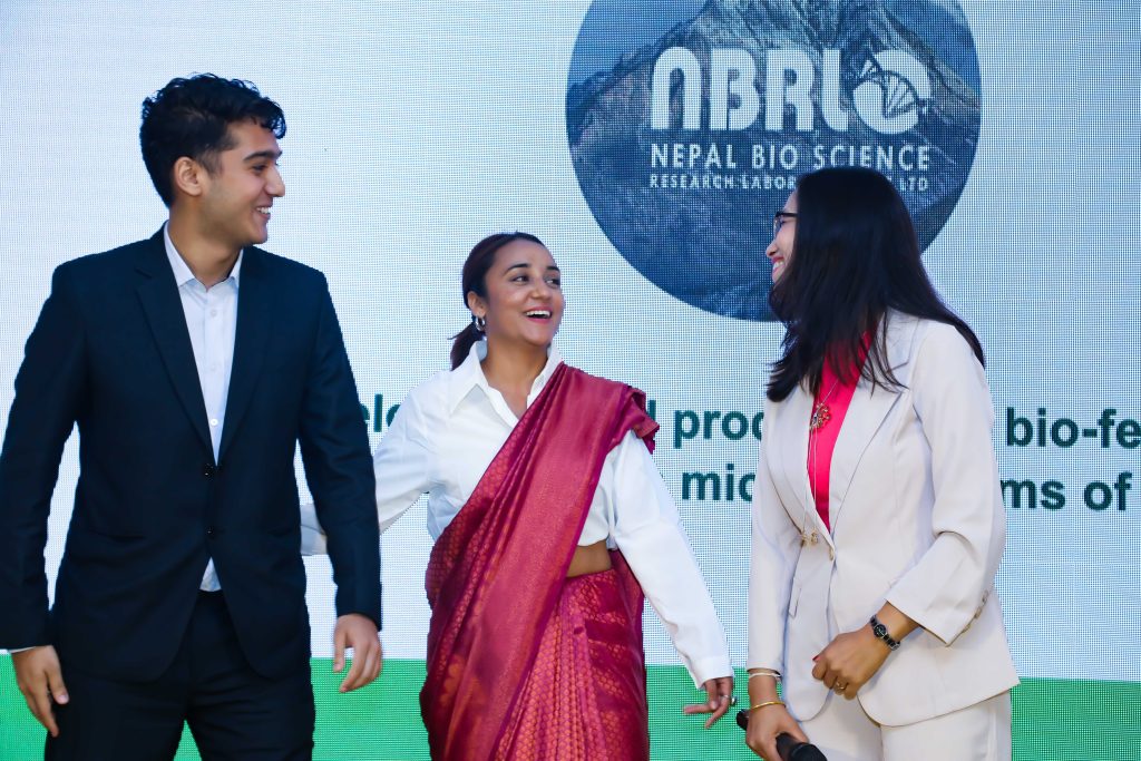 Three people laugh together while standing in front of a backdrop that reads "Nepal bio science"