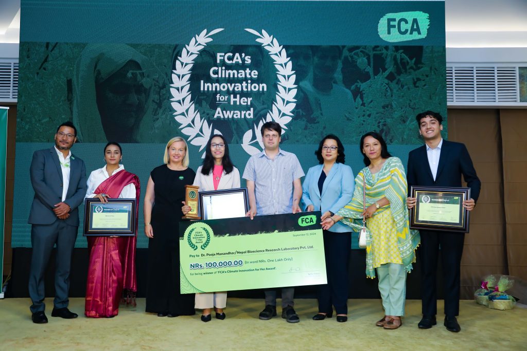 Eight people pose for a photo. Some of them are holding framed certificates. The three central people are holding a large cheque for 100,000.00 Nepali Rupees. A backdrop reads "FCA's Climate Innovation for Her Award"