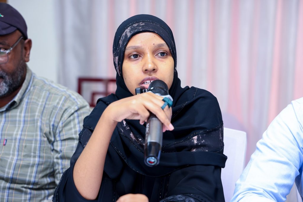 A woman in a hijab sits at a table and speaks into a microphone