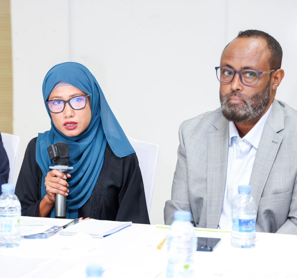 A woman in a hijab speaks into a microphone while seated at a desk. A man sits next to her.