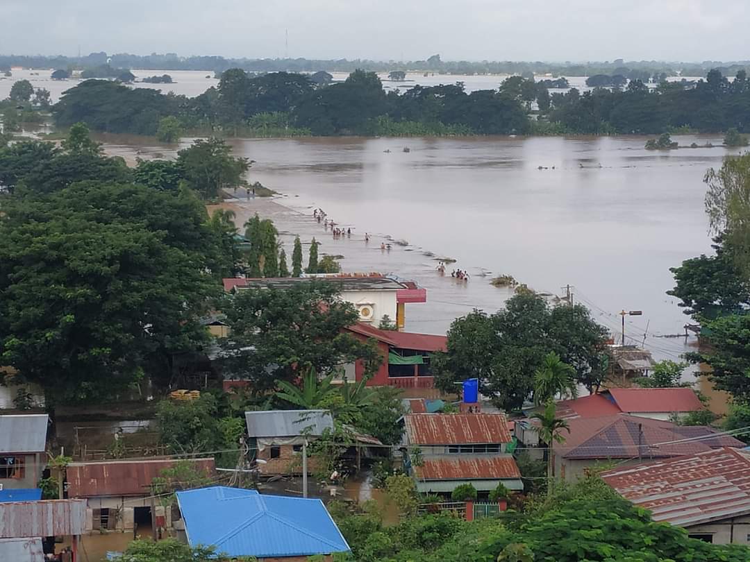 A view from a hilltop of a flooded village and fields. People in the distance trudge through floodwaters.