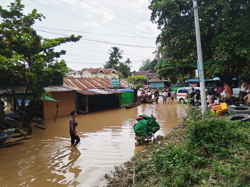 A flooded street. People walk through the waters dragging boats. Simple shacks line the street and are full of water