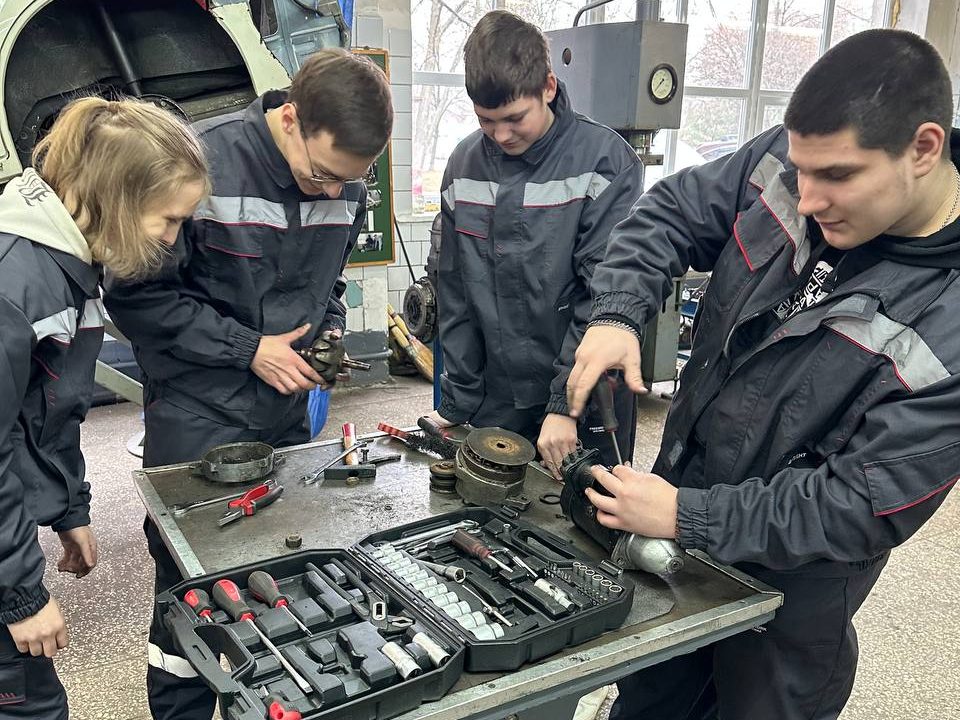 Four people in workclothes stand around a workbench which has a number of tools on it. They are all busy working.