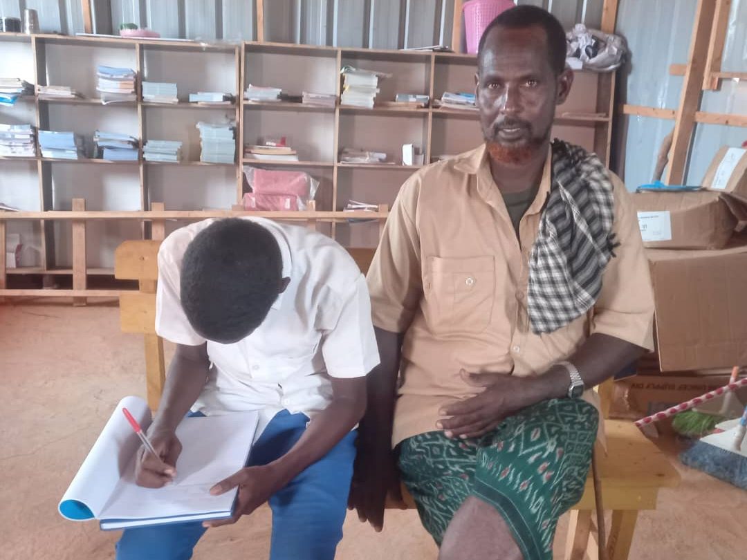 A man sits on a bench in an office and looks at the camera. Next to him a teenaged boy is busy writing in an exercise book.