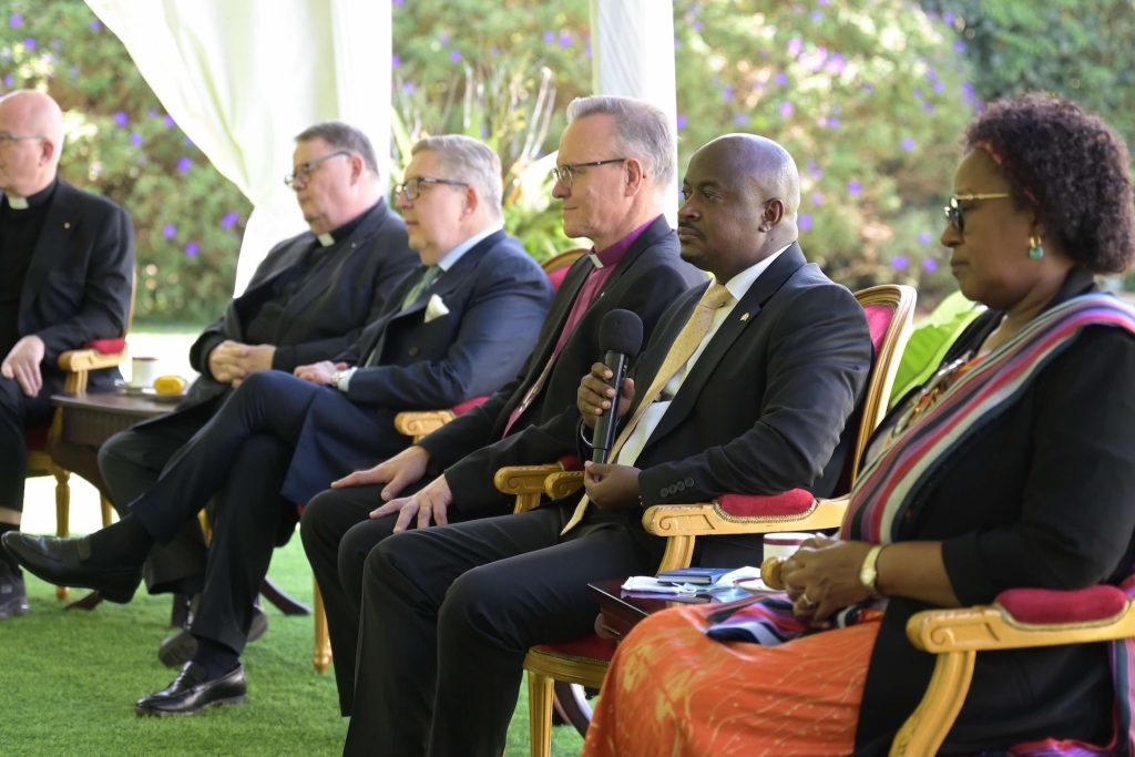 Men and women in business attire or clerical collars sit in ornamental chairs on grass under a marquee. One is holding a microphone