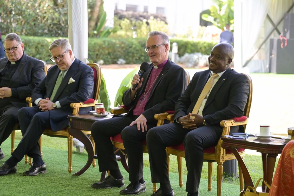 Four men in business suits or clerical collars sit in ornamental chairs on grass under a marquee. One is talking into a microphone