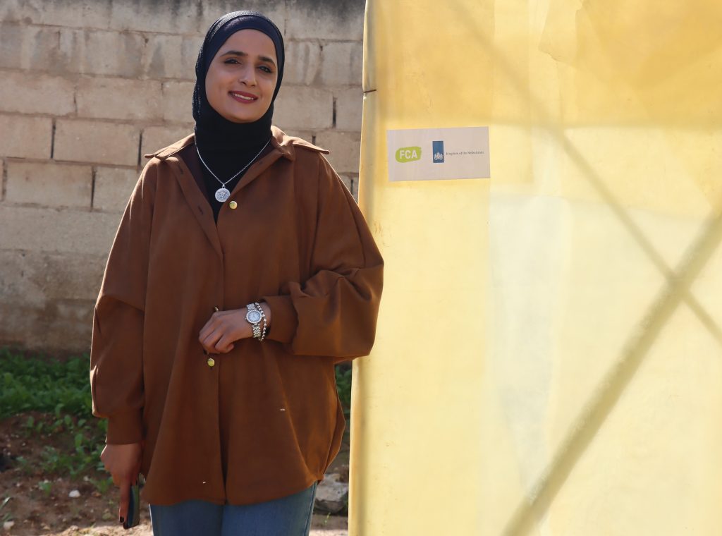 A woman stands outside in a field. Beside her a yellow canvas structure bears a sticker with the logo of FCA and the Kingdom of the Netherlands