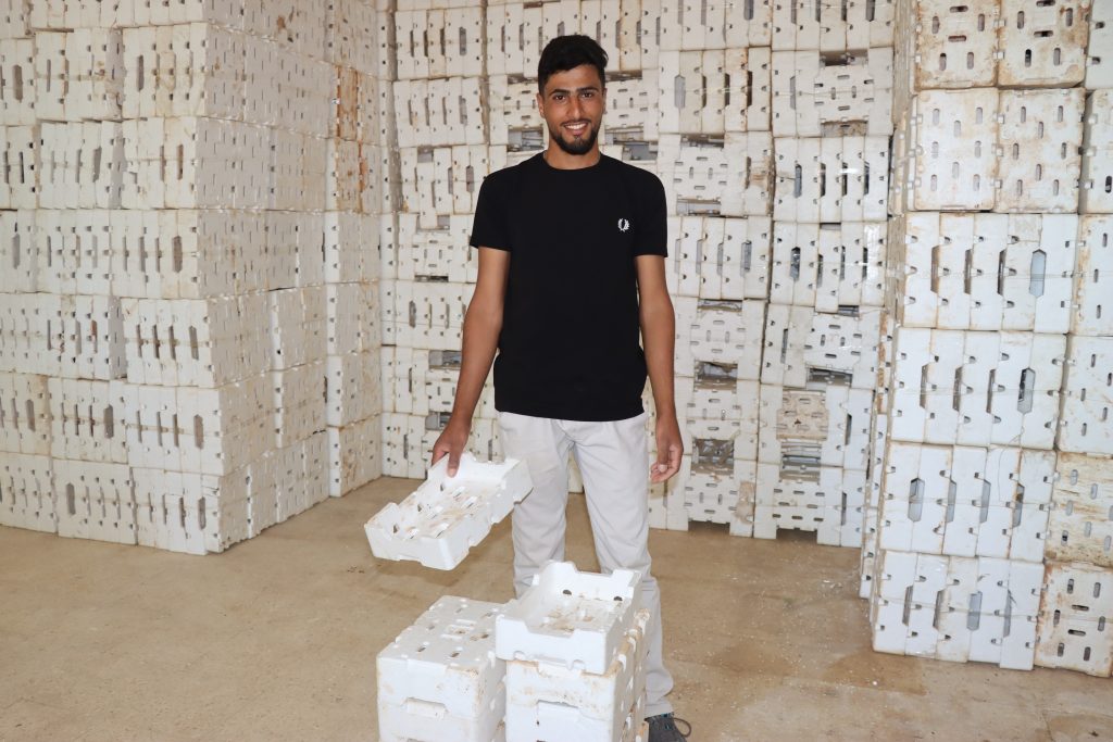 A smiling man stands with a pile of boxes in front of him. Behind him the same boxes are piled up to the ceiling.