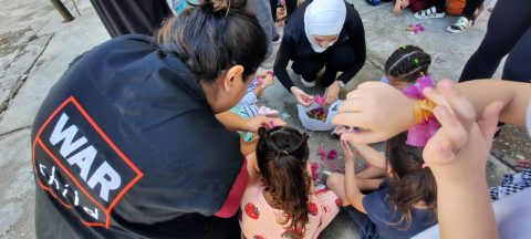 A person in a War Child jacket bends down to help children make bracelets in an outdoor setting