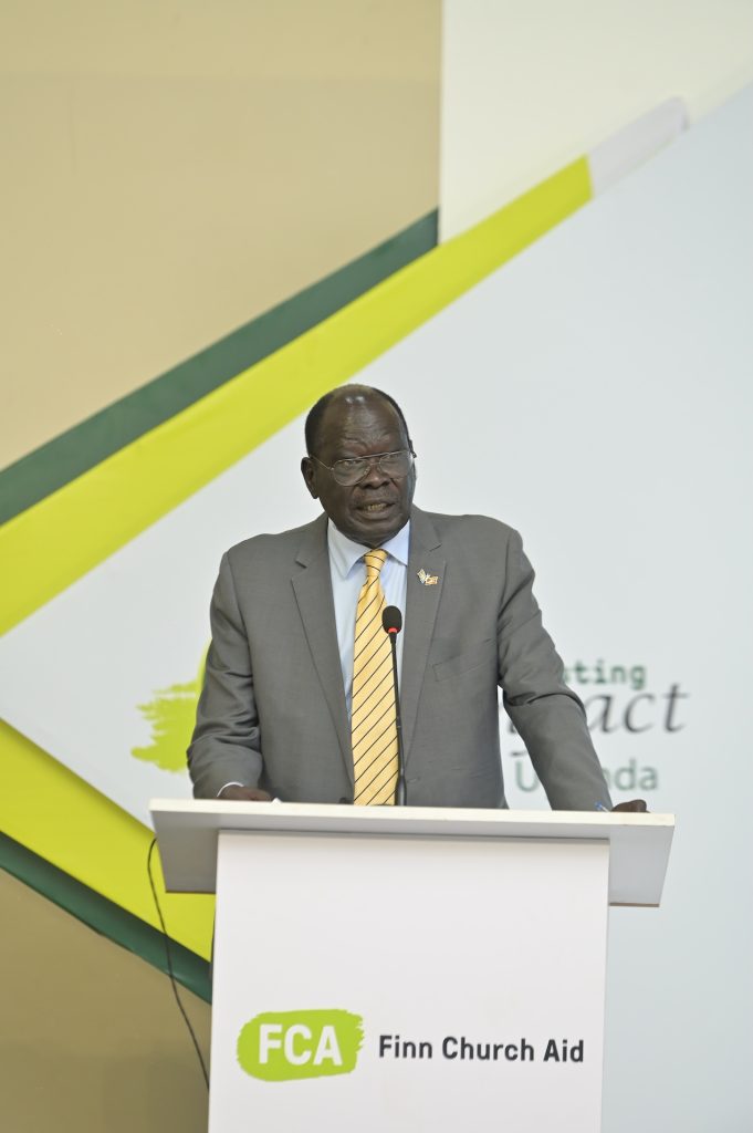 A man in a suit speaks into a microphone at a podium. The podium bears the FCA logo.