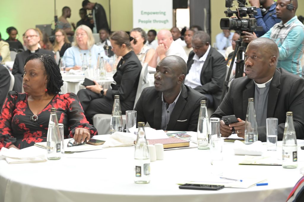 Three people sit at a round dining table in a crowded conference room. In the background there are more seated guests and a camera team filming.