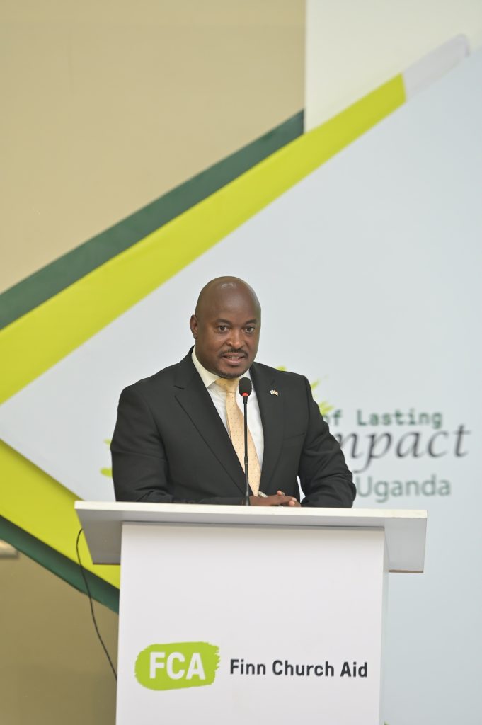 A man in a suit speaks into a microphone at a podium. The podium bears the FCA logo.