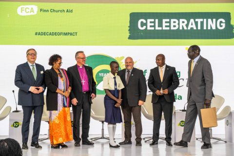 A line of people stand on a stage and pose for a photo. In the background there is the FCA logo and the hashtag #ADecadeOfImpact next to a large 'Celebrating' in green.