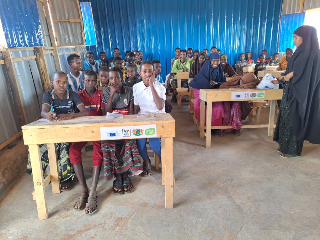 A classroom of children sitting at desks. They are laughing and looking at the camera. The desks bear the logos of FCA, Gredo and the EU