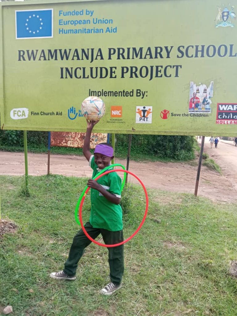 A smiling boy in an FCA green shirt holds up a football while standing in front of a large sign that reads Rwamwanja Primary School Include Project