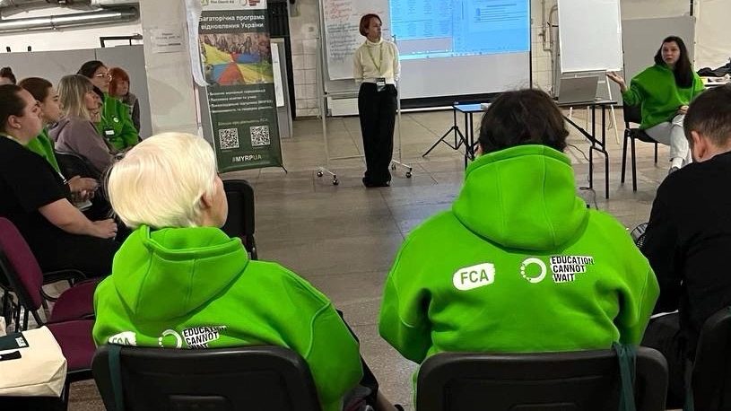 Two people in green hoodies bearing the logos of FCA and Education Cannot Wait are sitting in chairs in a circle of chairs in a conference room. Two people are giving a presentation to them. 