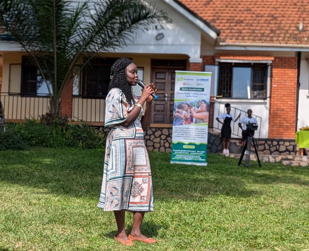 A woman with a microphone stands on a lawn and makes a speech. In the background is a banner with FCA and UNHCR logos.