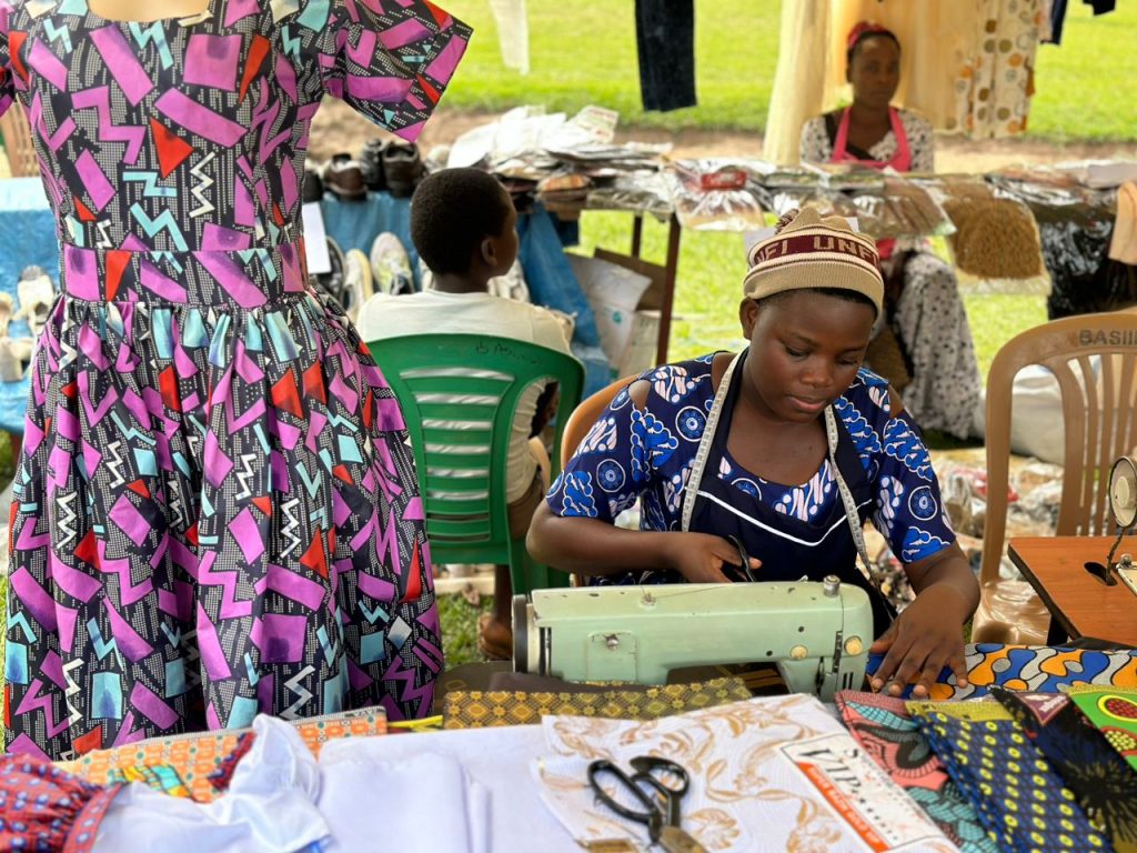 A woman sitting at a sewing machine at a market. She has a mannequin next to her.