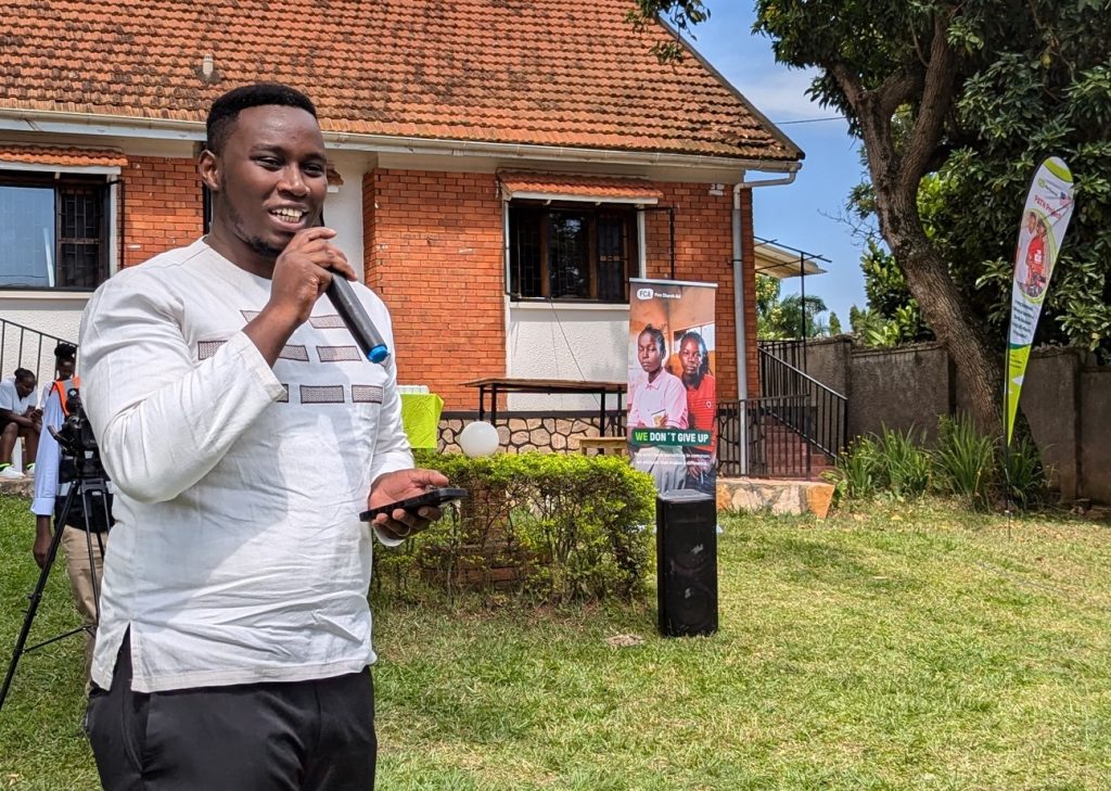 A man with a microphone stands on a lawn and makes a speech. In the background are several banners with FCA and UNHCR logos.