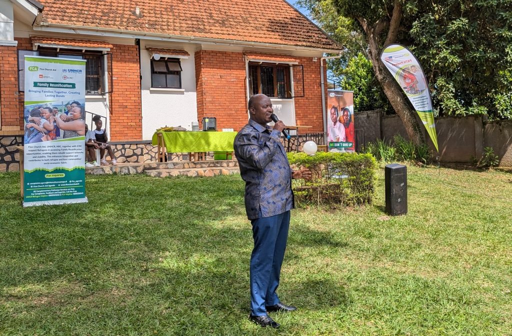 A man with a microphone stands on a lawn and makes a speech. In the background are several banners with FCA and UNHCR logos.