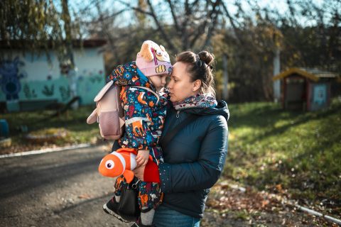 En kvinna håller ett litet barn i sin famn. I bakgrunden syns ett parklandskap.