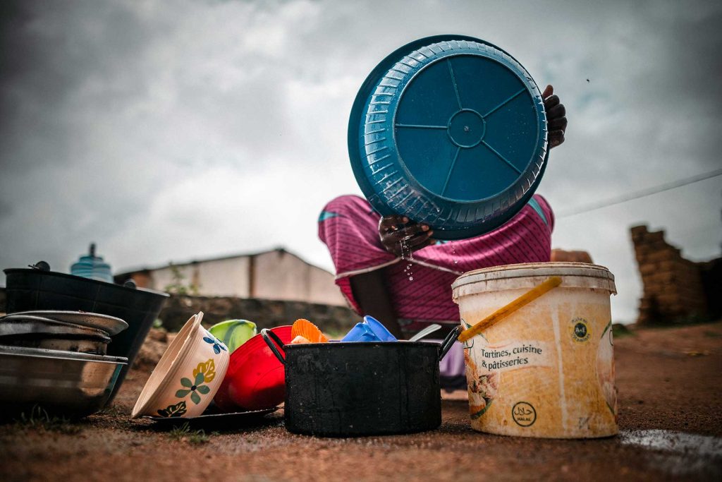 A person crouching on the ground holds up a large plastic bowl, obscuring their face.