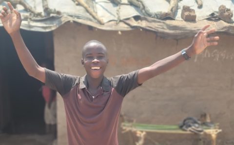 A young boy standing in front of a mud hut with his hands up in celebration and a big smile on his face.