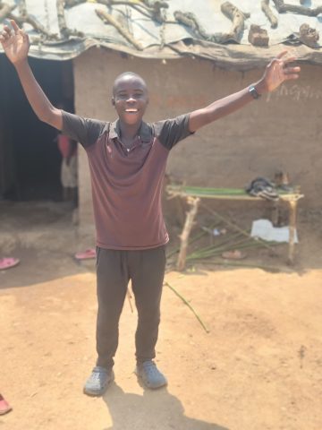 A young boy standing in front of a mud hut with his hands up in celebration and a big smile on his face.