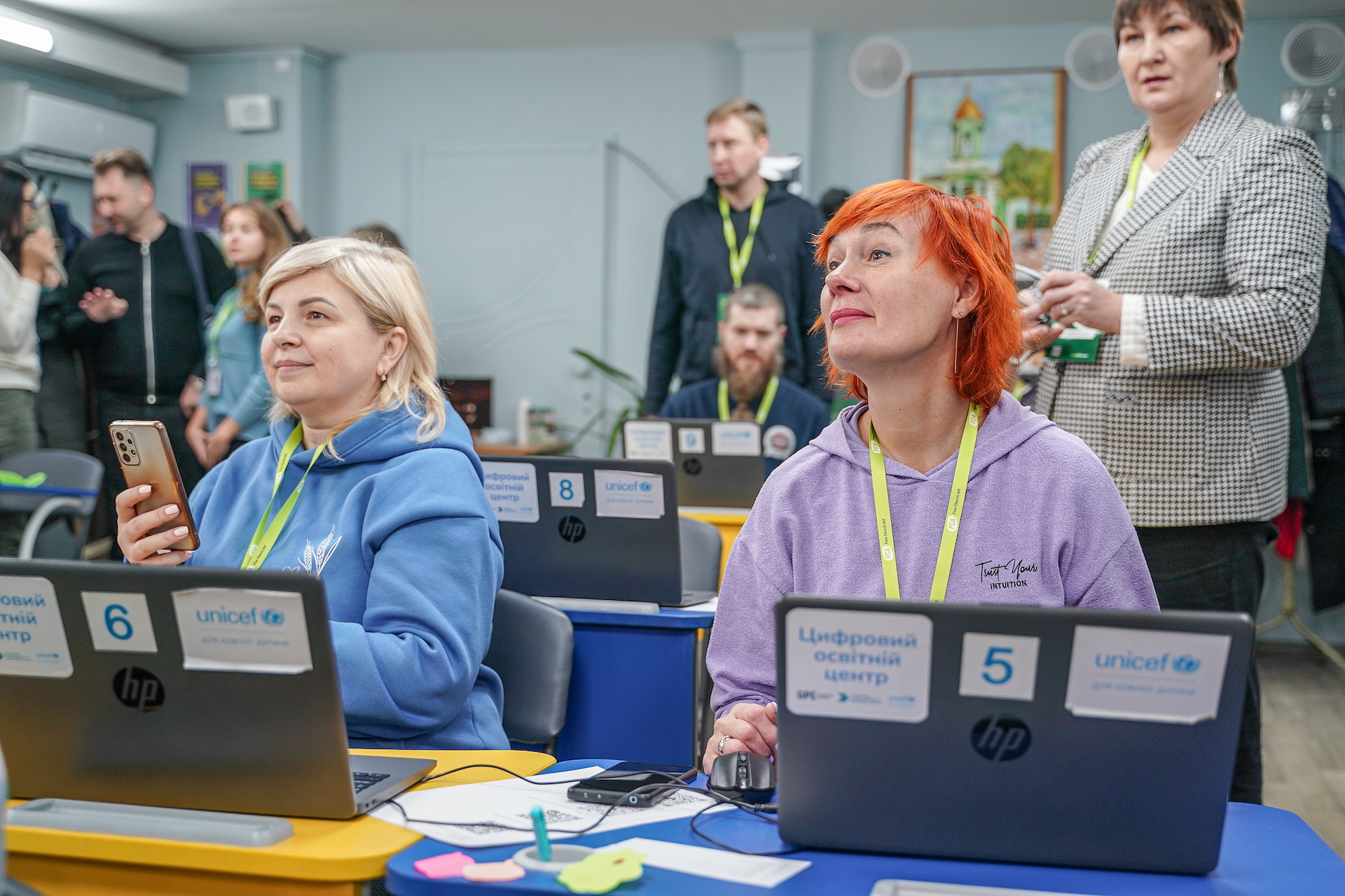 Two women at a desk with laptops in a classroom look up to listen. One is holding a mobile phone. 