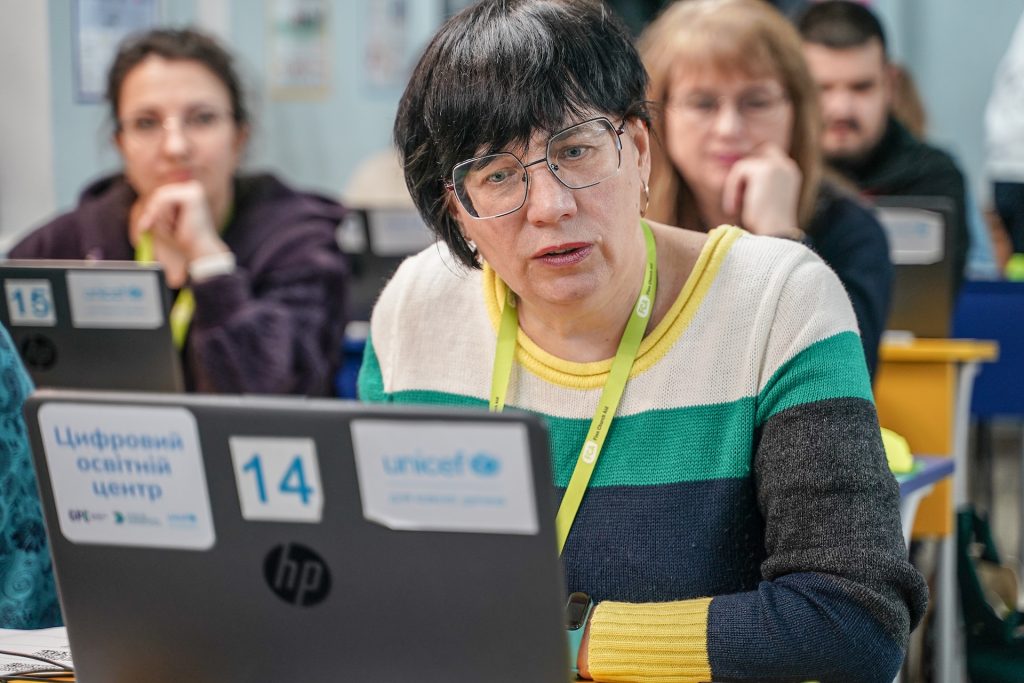 A woman sits at a laptop and looks at the screen. Behind her there are other people at desks in a classroom environment. 