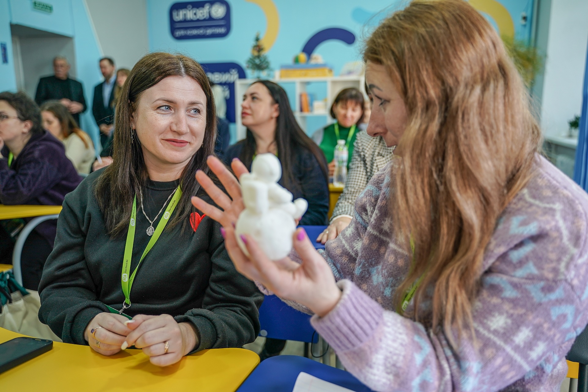 Two women sit at a desk in a classroom and talk. 